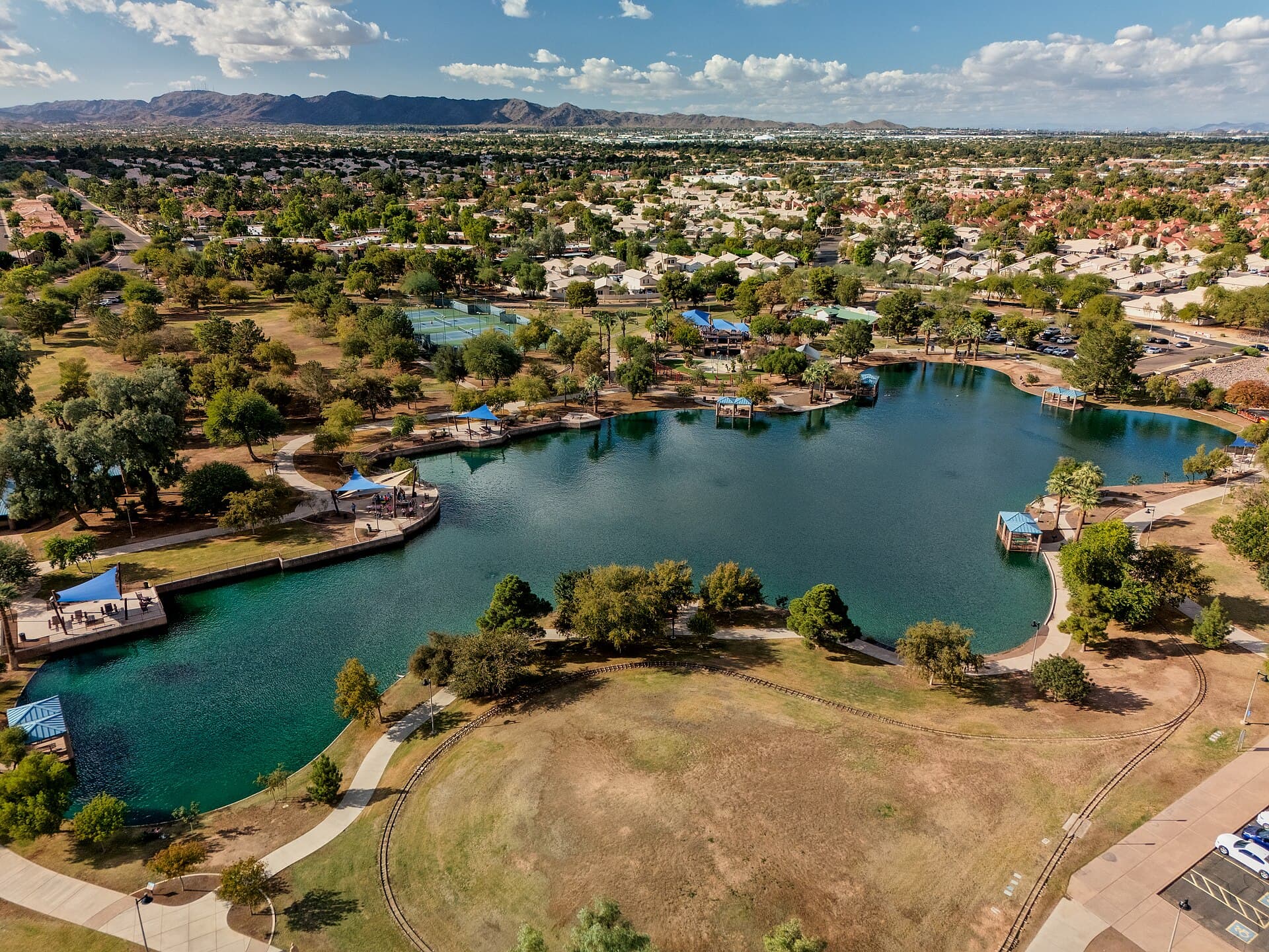 Aerial view of Desert Breeze Lake in Chandler, Arizona, with ramadas and walking paths, surrounding neighborhoods and a mountain range on the horizon.