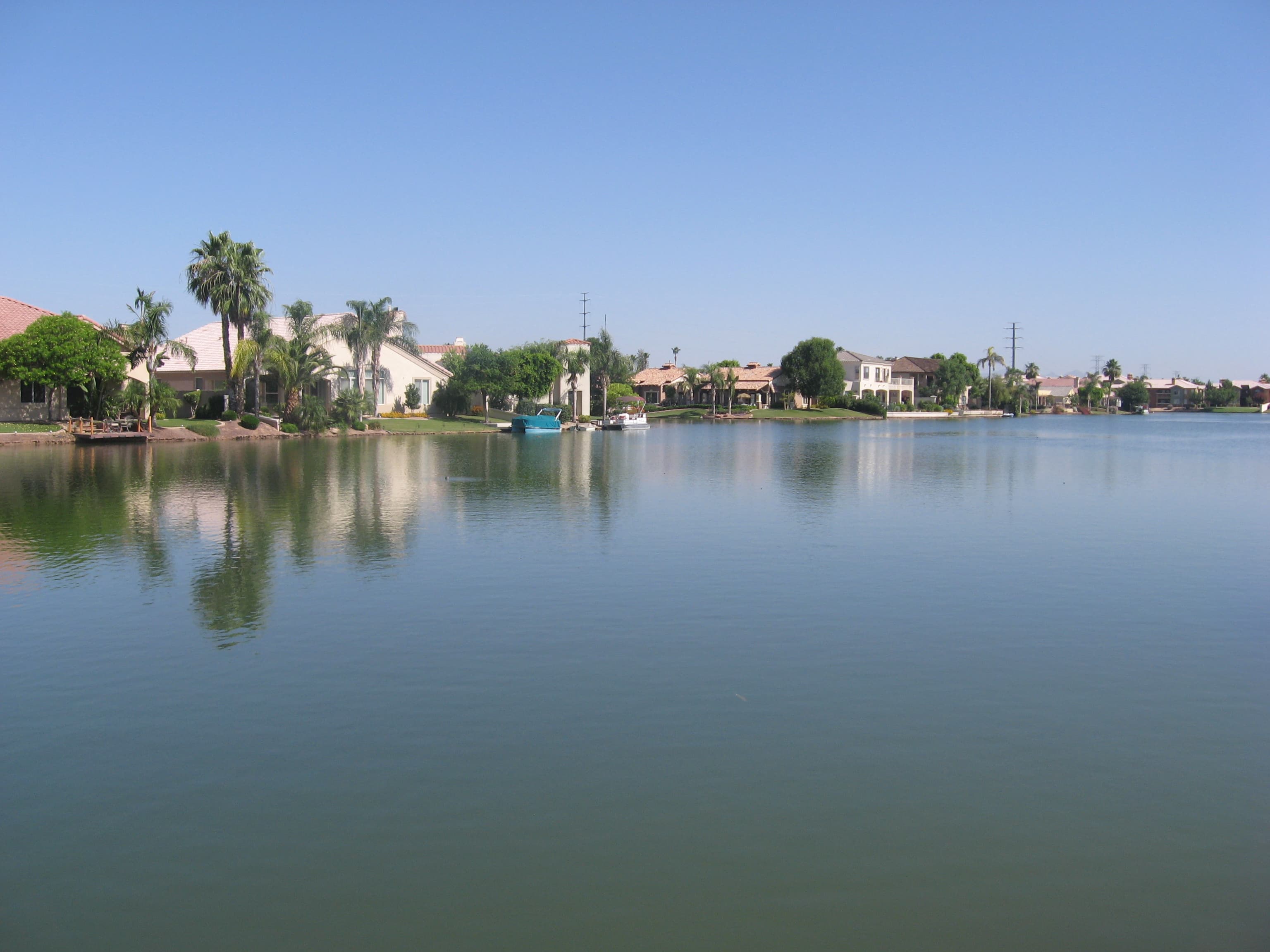 A calm residential lake in the Val Vista Lakes community in Gilbert, Arizona, with palm trees and homes along the far shore under a clear sky.